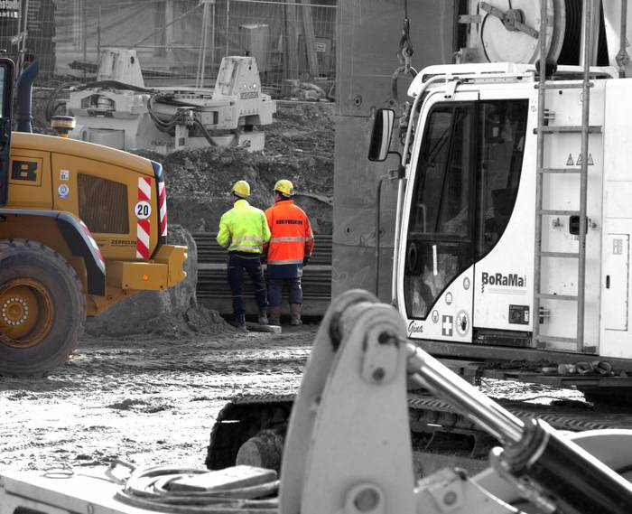 Worker stand close to and facing away from construction vehicles.