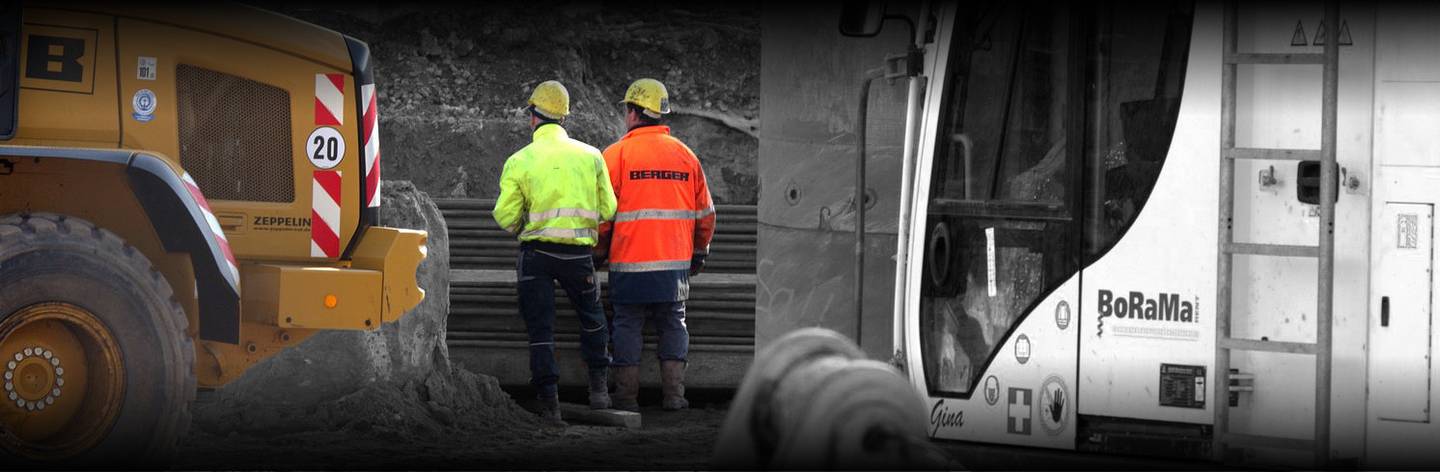 Worker stand close to and facing away from construction vehicles.